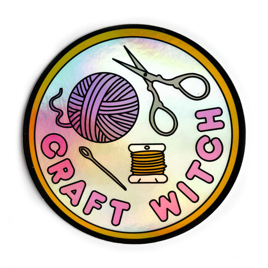 A circular holographic sticker. It has a yellow borer with a light blue background and hot pink text that reads "Craft Witch". Above the words a needle, thread, yarn ball, and scissors are depicted.