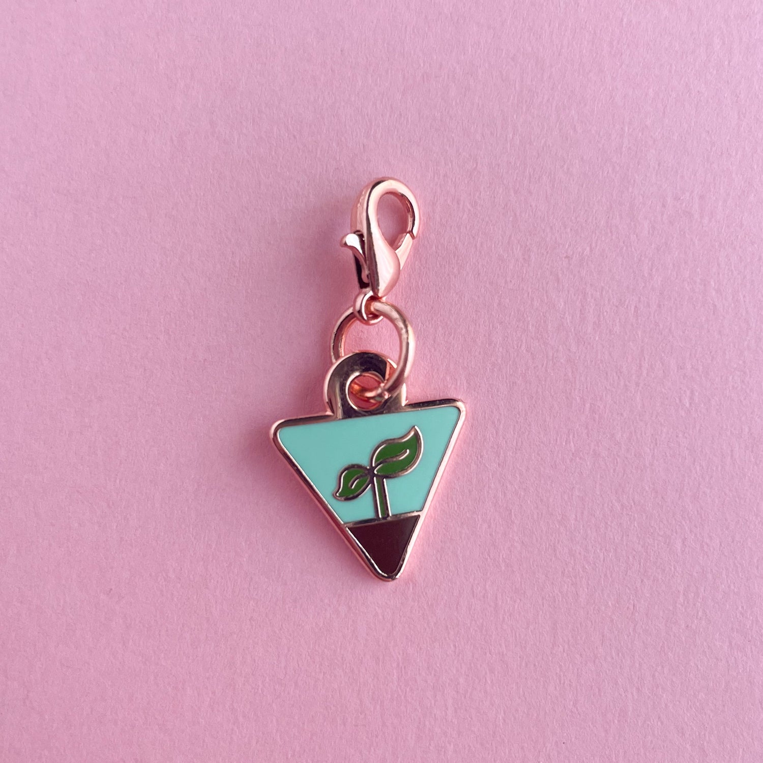 A charm in the shape of an upside down triangle with a small plant sprout growing out of some dirt on it.  The charm is on a pink background.