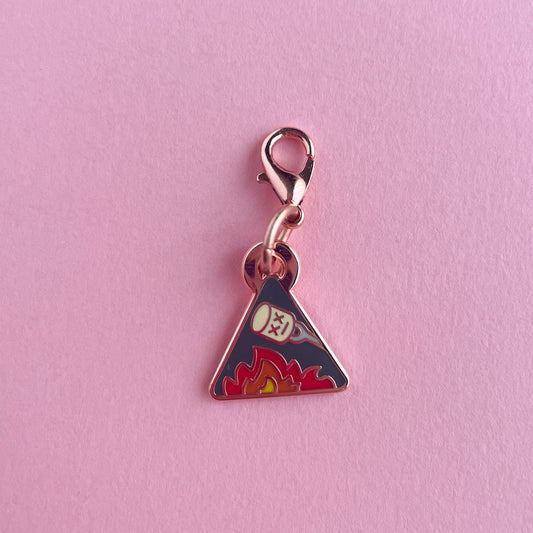A triangle shaped charm with a marshmallow roasting over a flame. The charm is on a pink background.