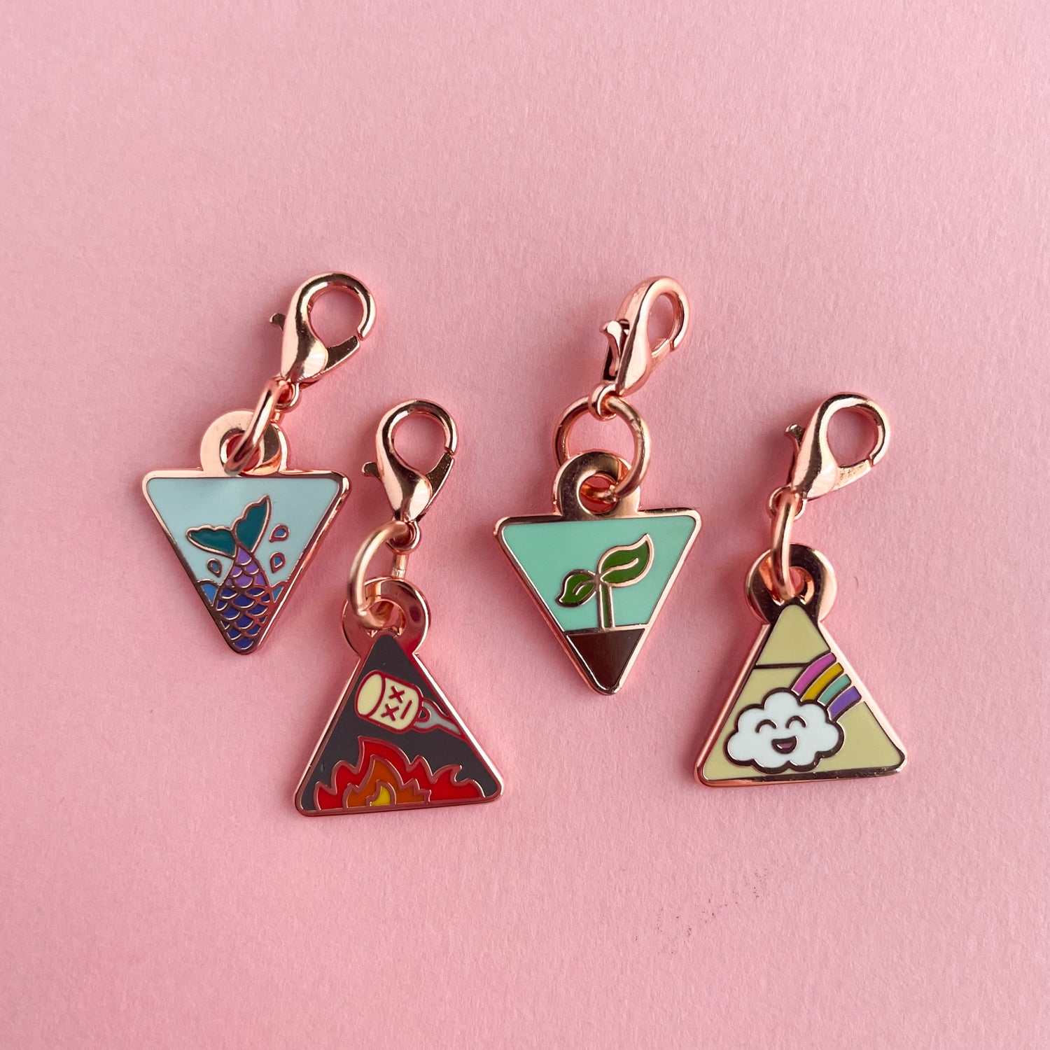 Four triangle shaped charms with lobster claw clasps. There is a mermaid tail charm, a fire with a marshmallow charm, a plant sprouting charm, and a happy rainbow cloud charm.  The charms are on a pink background