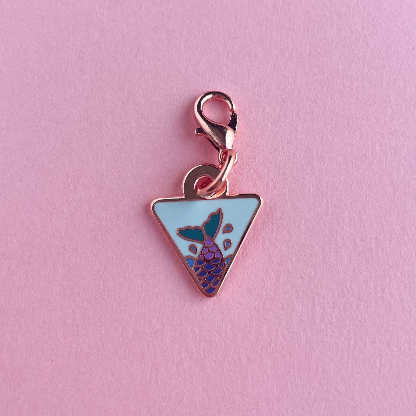 An upside down triangle charm with a lavender mermaid tail on it. The charm is on a pink background.