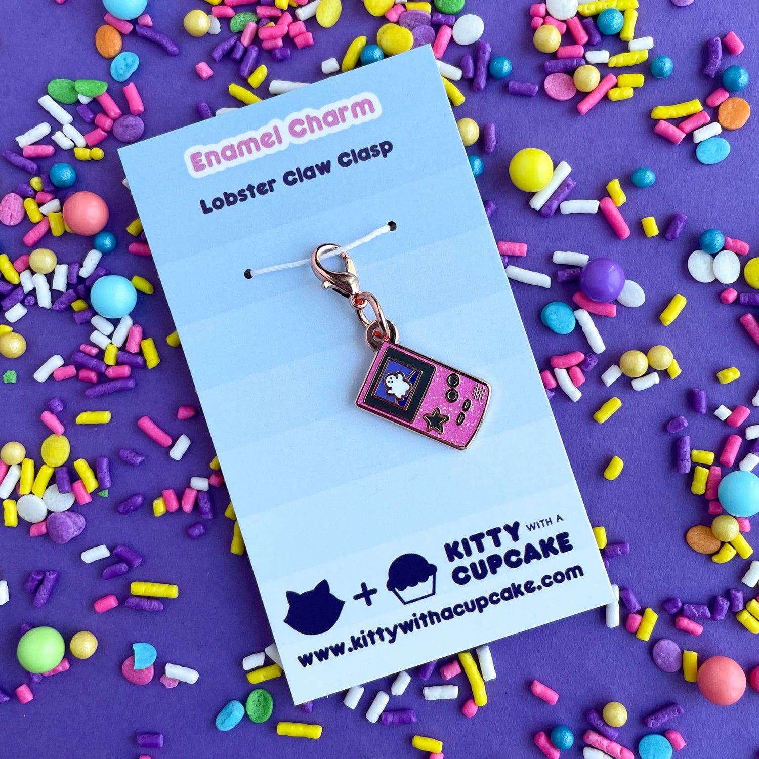 A lobster claw clasp charm that is the shape of a pink glitter Game Boy with a star button and a cute ghost on the screen. The charm is packaged on a backing card that reads "Enamel Charm - Lobster Claw Clasp". It is on a purple background that is covered in pastel sprinkles.