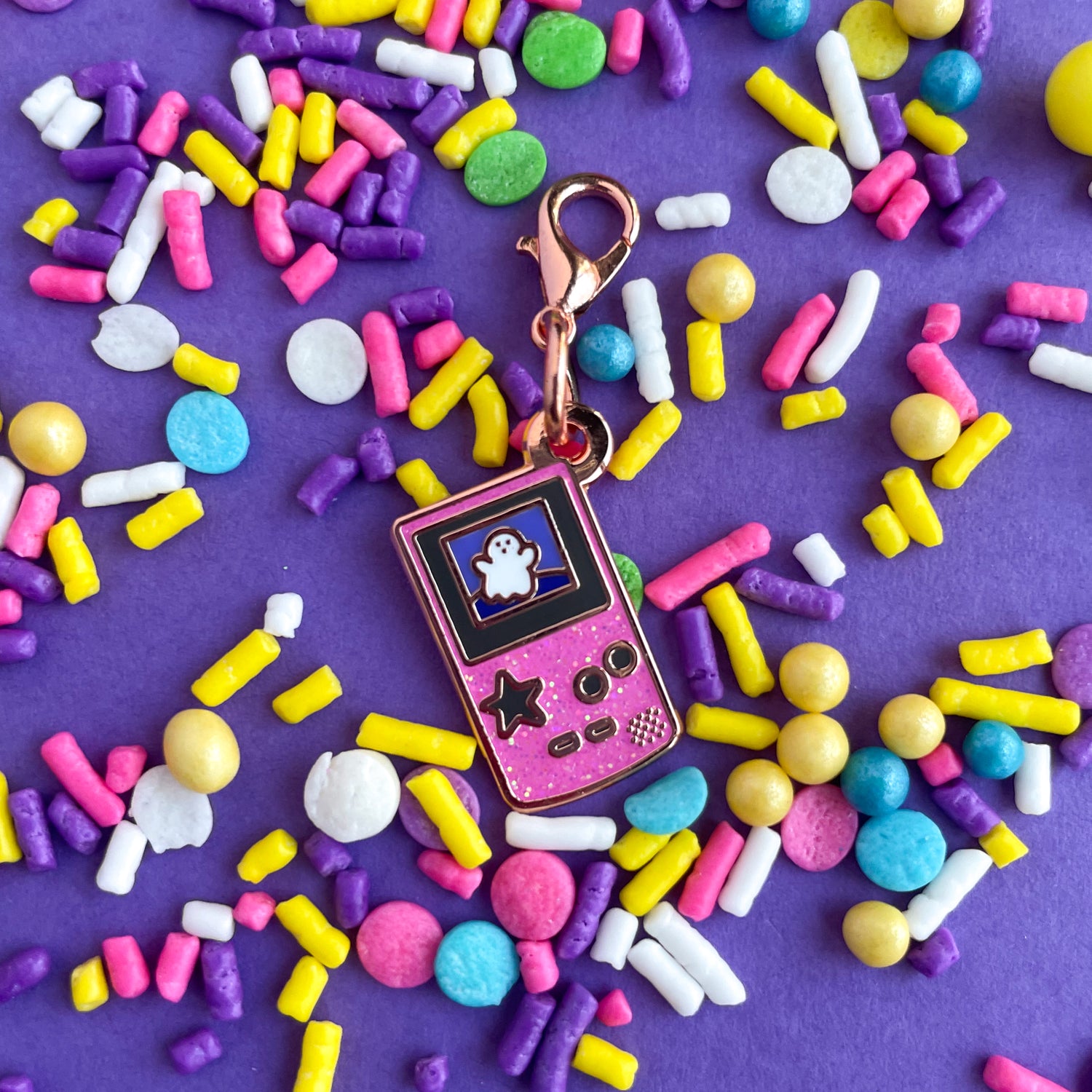 A lobster claw clasp charm that is the shape of a pink glitter Game Boy with a star button and a cute ghost on the screen. It is on a purple background covered in pastel sprinkles.