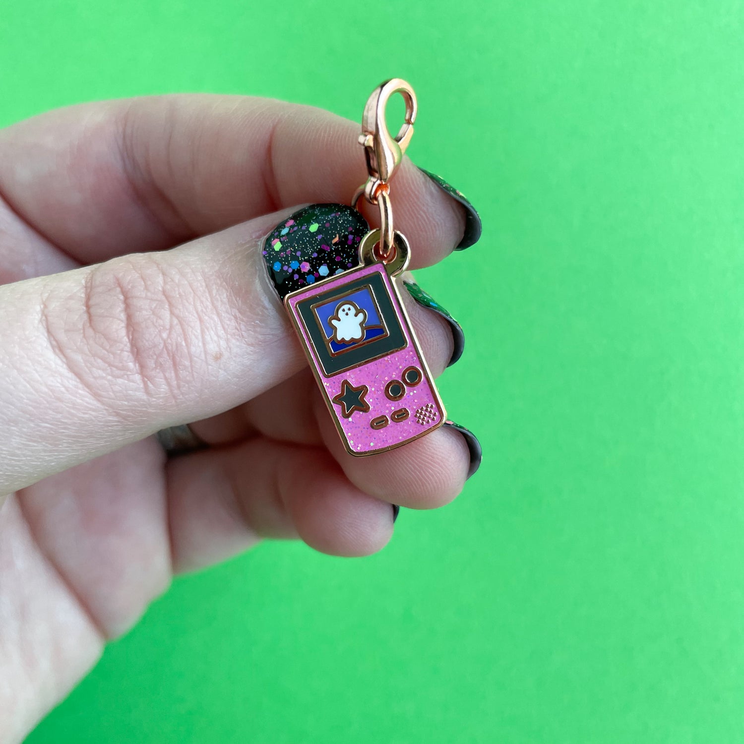A hand with black and confetti nails holding a lobster claw clasp charm that is the shape of a pink glitter Game Boy with a star button and a cute ghost on the screen. This is on a green paper background.