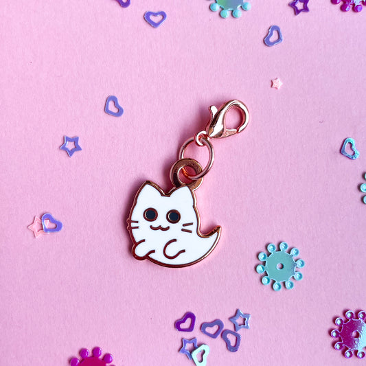A charm of a cat ghost in rose gold colored metal on a pink paper background with confetti around it. 