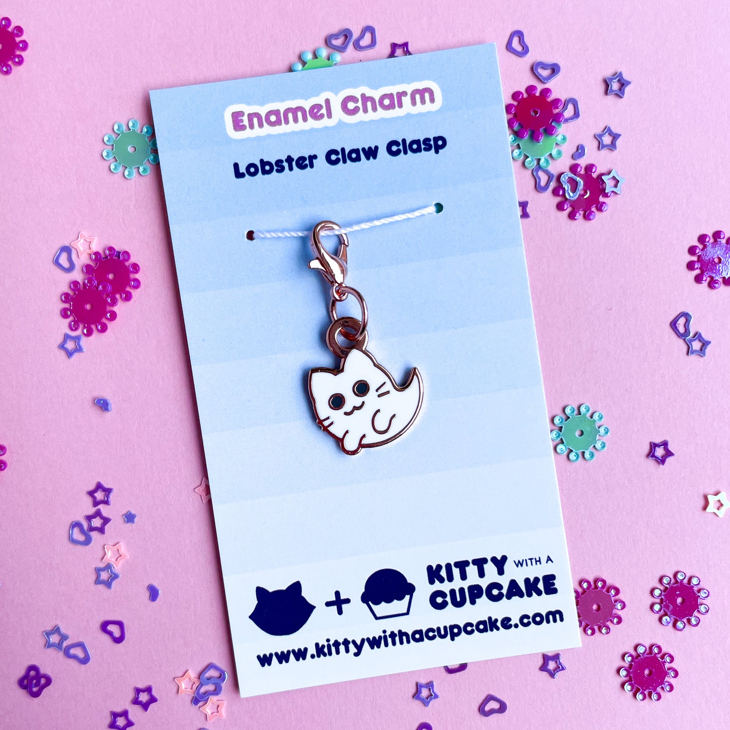 A cat ghost charm on a blue card that reads "Enamel Charm" on a pink paper background that is covered in confetti. 