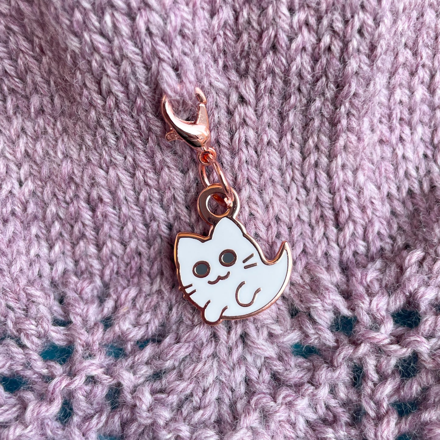 A kitty ghost charm attached to a piece of knitting that is mauve stockinette stitch going into lace. 
