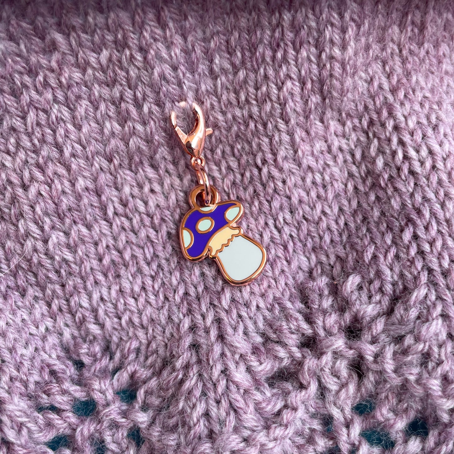 A rose gold colored mushroom shaped lobster claw clasp charm attached to a piece of mauve knitting. 