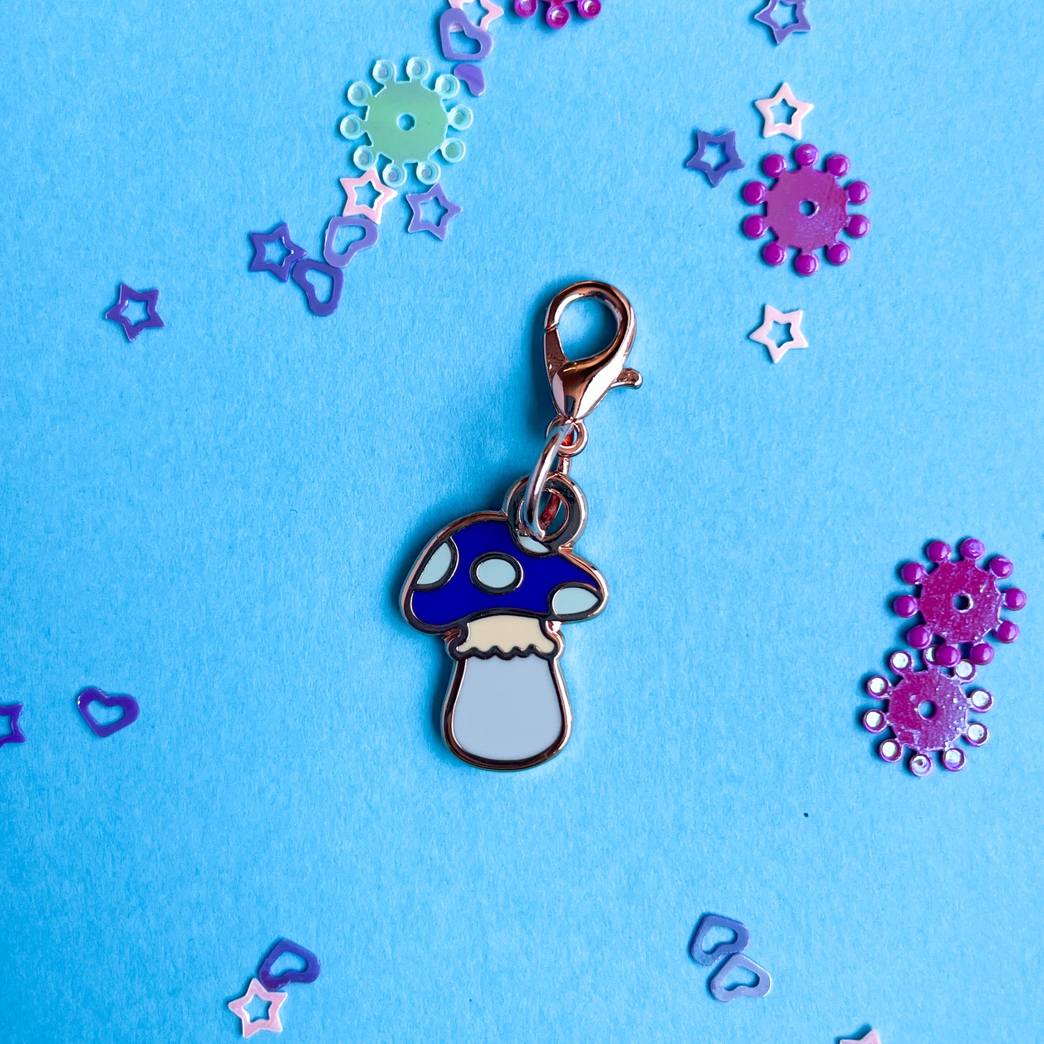 A mushroom shaped lobster claw clasp charm with a purple head and blue spots on a blue background with purple confetti.