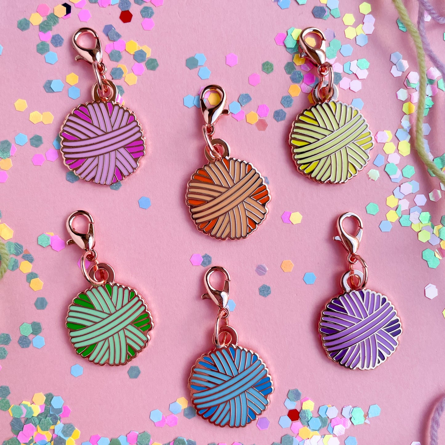 Six lobster claw clasp charms shaped like yarn balls in pink, orange, yellow, mint, blue, and purple on a pink background covered in confetti. 