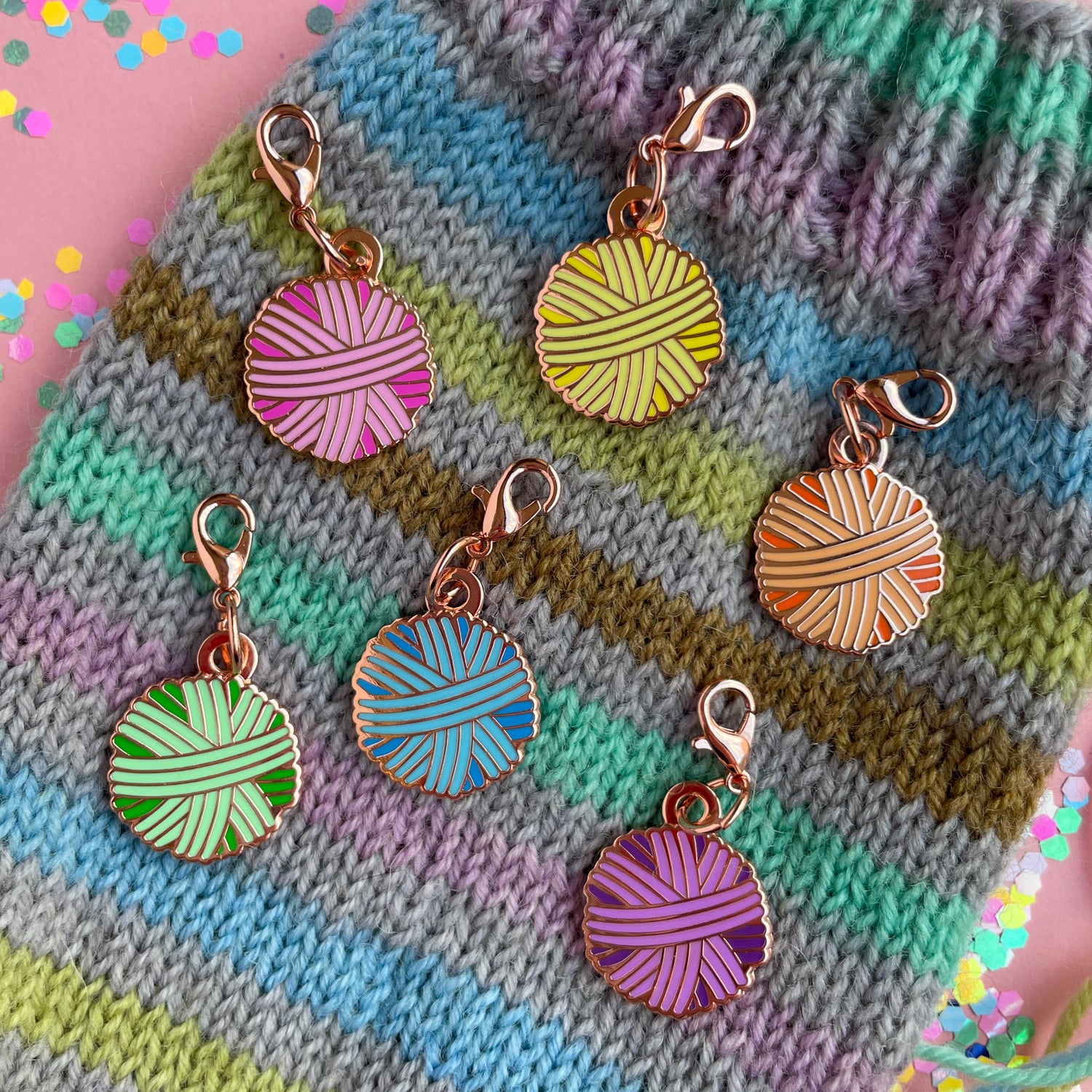 Six lobster claw clasp charms shaped like yarn balls in pink, orange, yellow, mint, blue, and purple on a grey and pastel rainbow striped hand knit sock. 