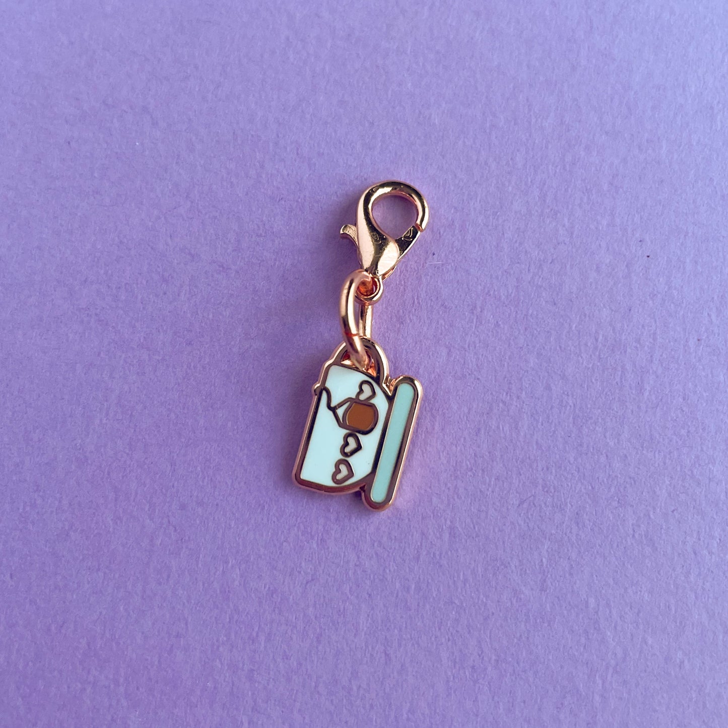 A charm on a lobster claw clasp that is shaped like a teacup with pink hearts on it and a teabag.  The charm is on a lavender background.