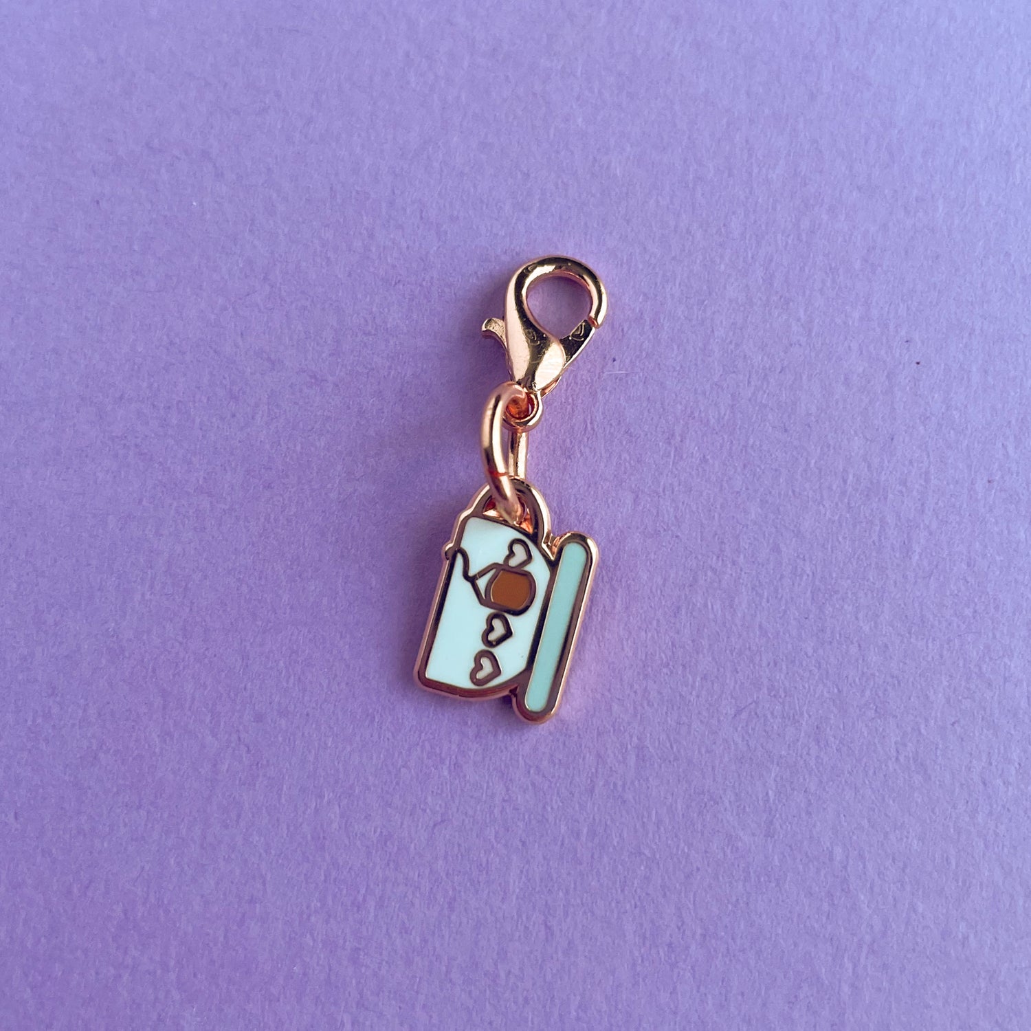A charm on a lobster claw clasp that is shaped like a teacup with pink hearts on it and a teabag.  The charm is on a lavender background.