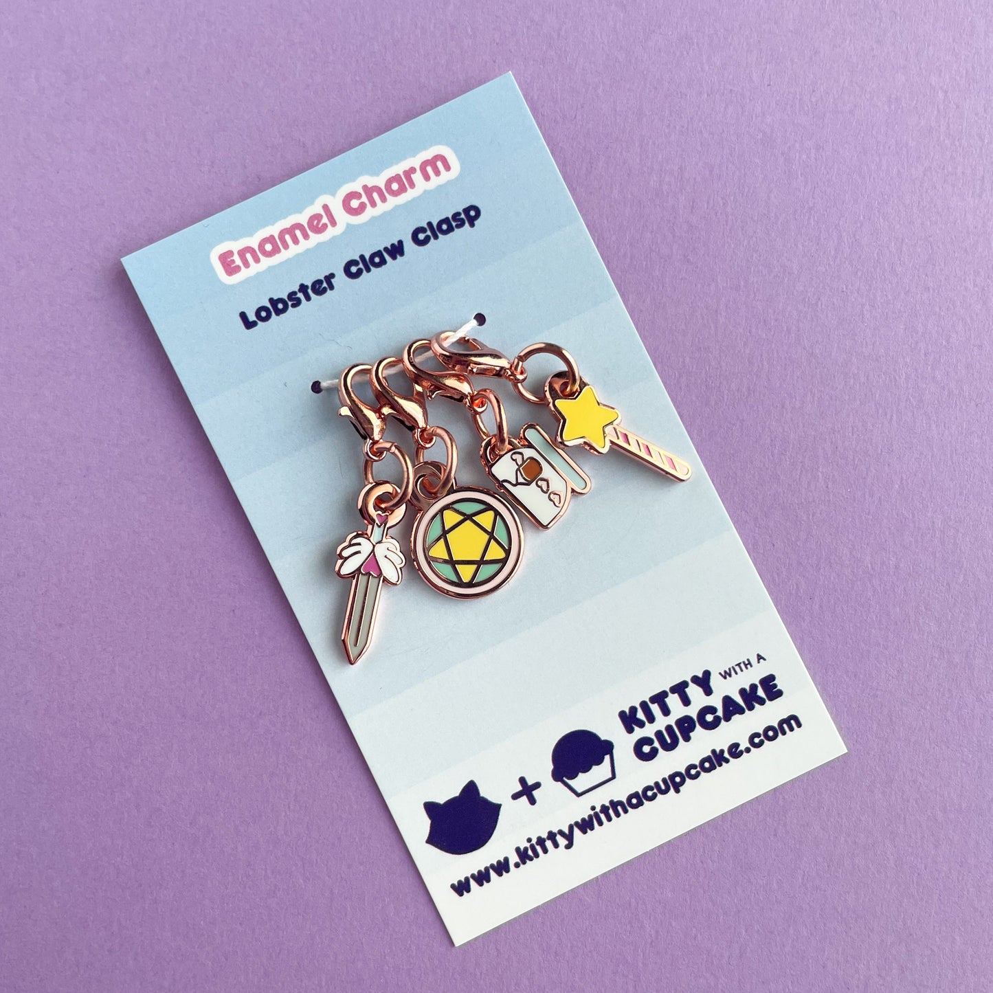 A collection of charms on a card on a lavender background. The charms are shaped like a sword, a teacup, a wand, and a pentacle circle.