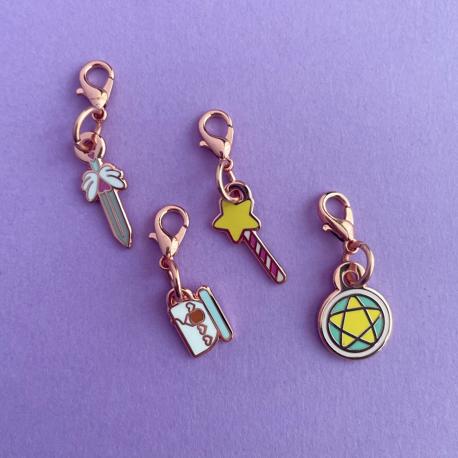 A collection of charms on a purple background. The charms are shaped like a sword, a teacup, a wand, and a pentacle circle.