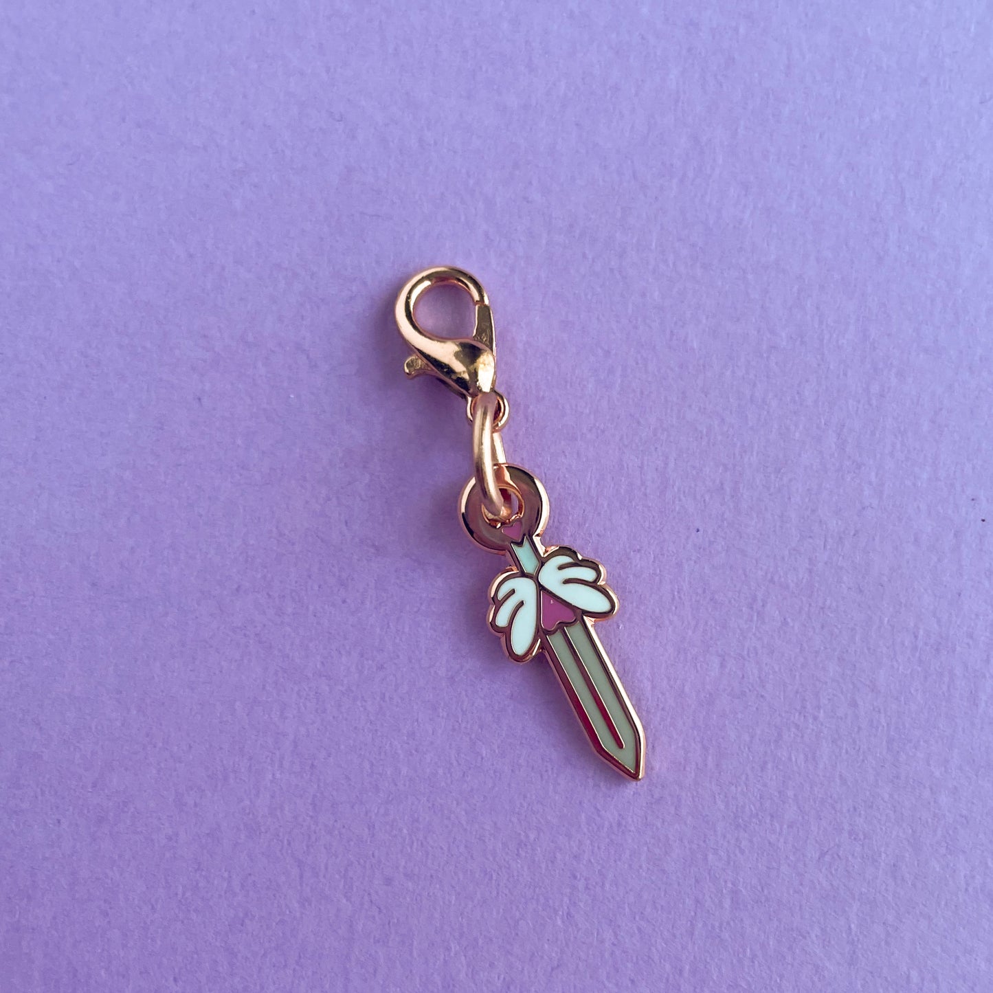 A charm with a lobster claw clasp in rose gold metal. The charm is shaped like a sword with heart and winged hilt. The charm is on a lavender background.