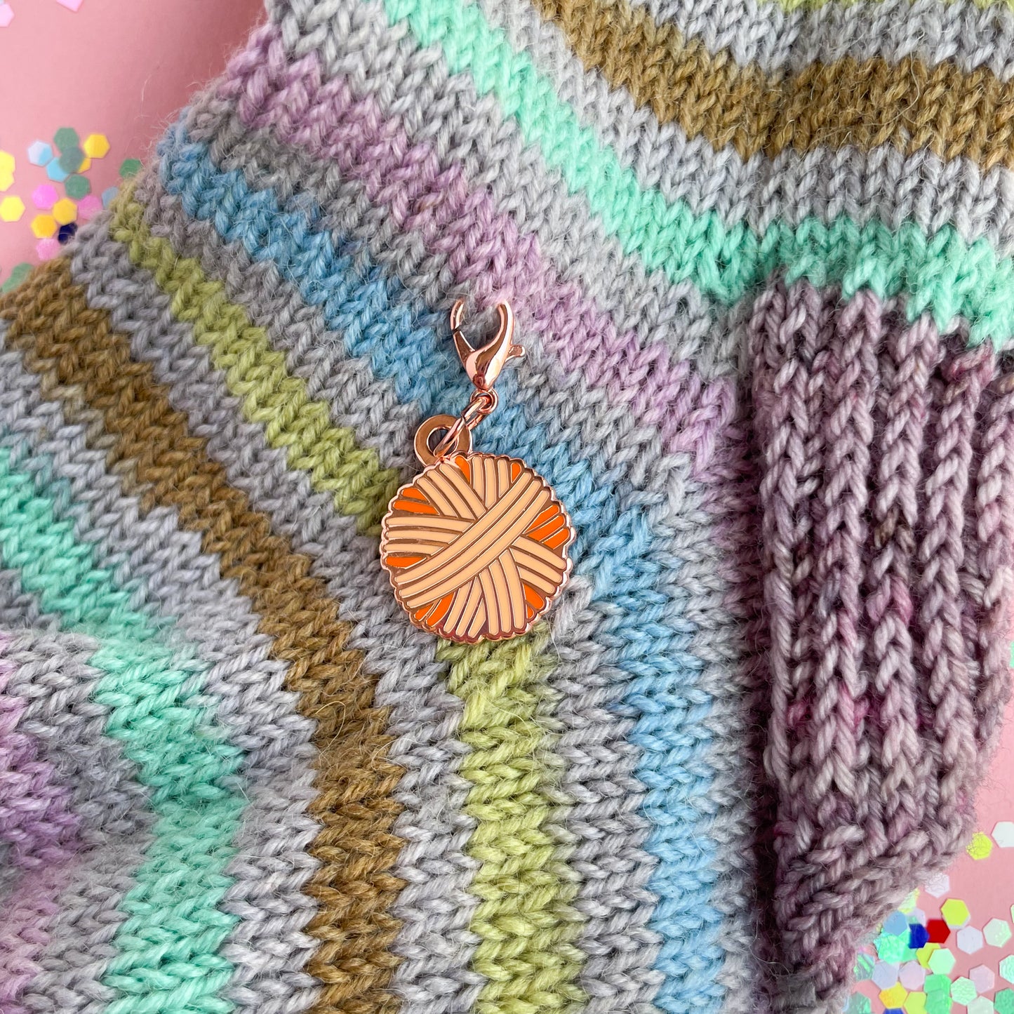 An orange yarn ball shaped charm on a hand knit sock that is grey with pastel rainbow stripes. 