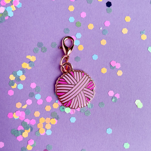 A pink yarn ball shaped charm with a lobster claw clasp on a lavender background with confetti. 