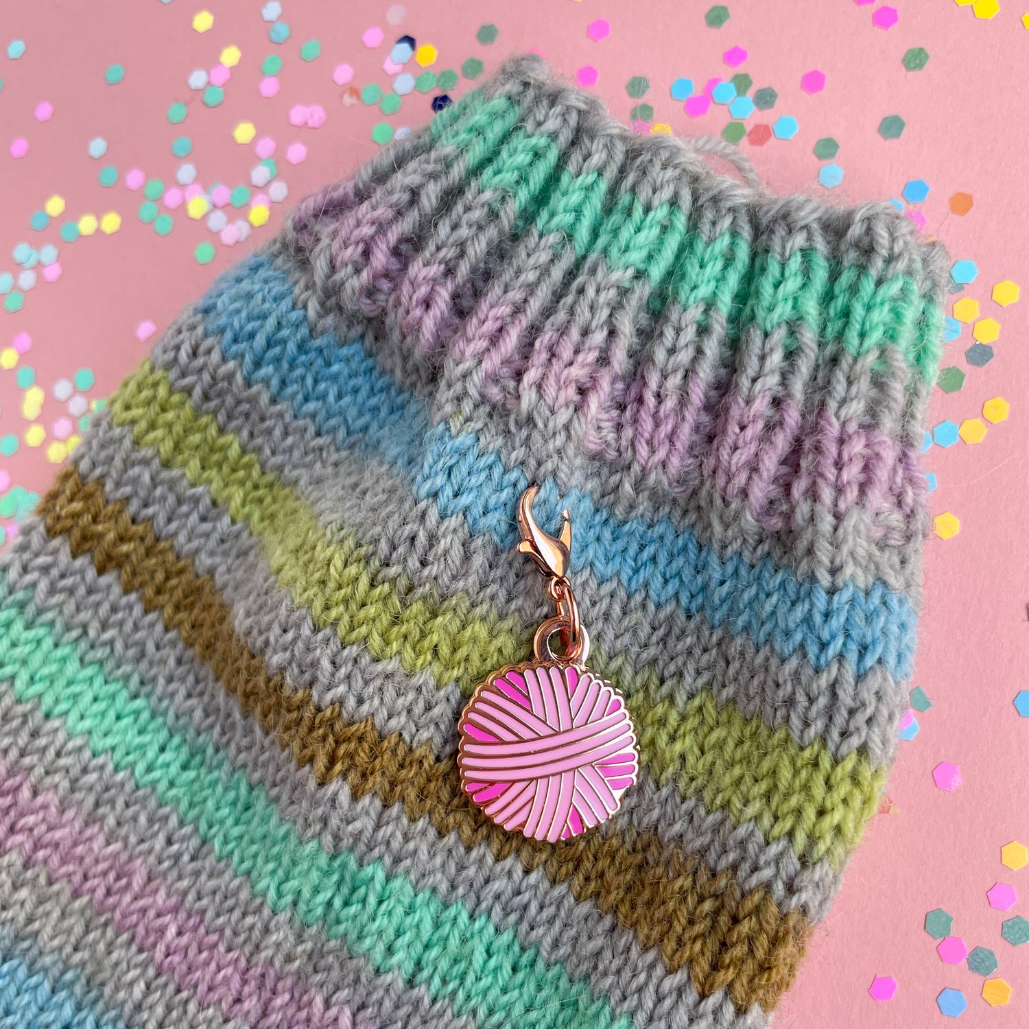 A pink yarn ball charm on a grey and pastel rainbow striped hand knit sock. 