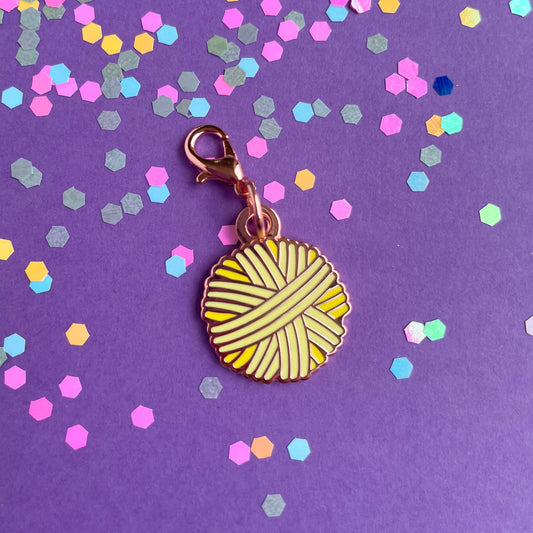 A yellow yarn ball shaped charm with a lobster claw clasp on a lavender background with confetti.