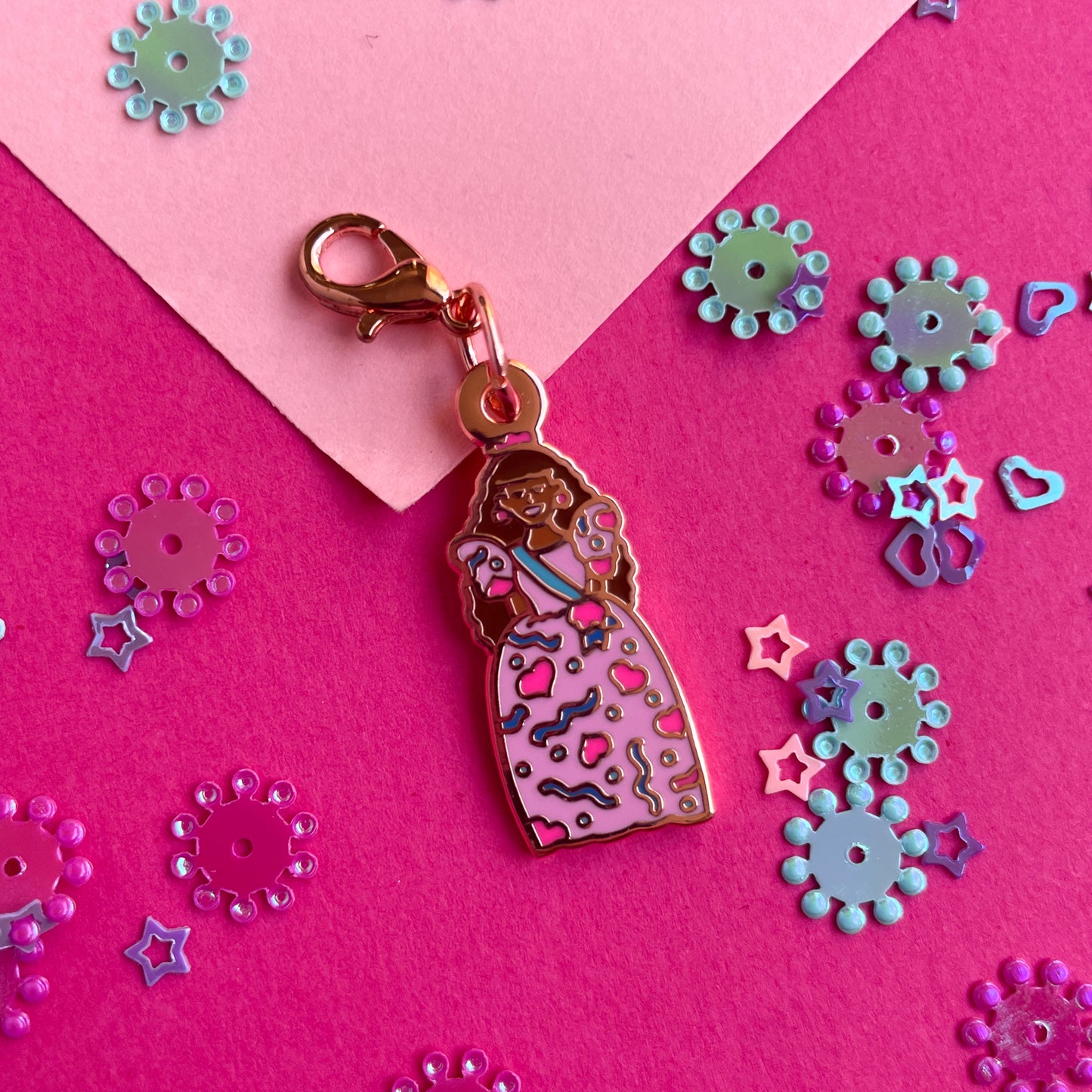An enamel charm with a lobster claw clasp shaped like a birthday barbie doll with caramel skin and brown hair on a pink background.
