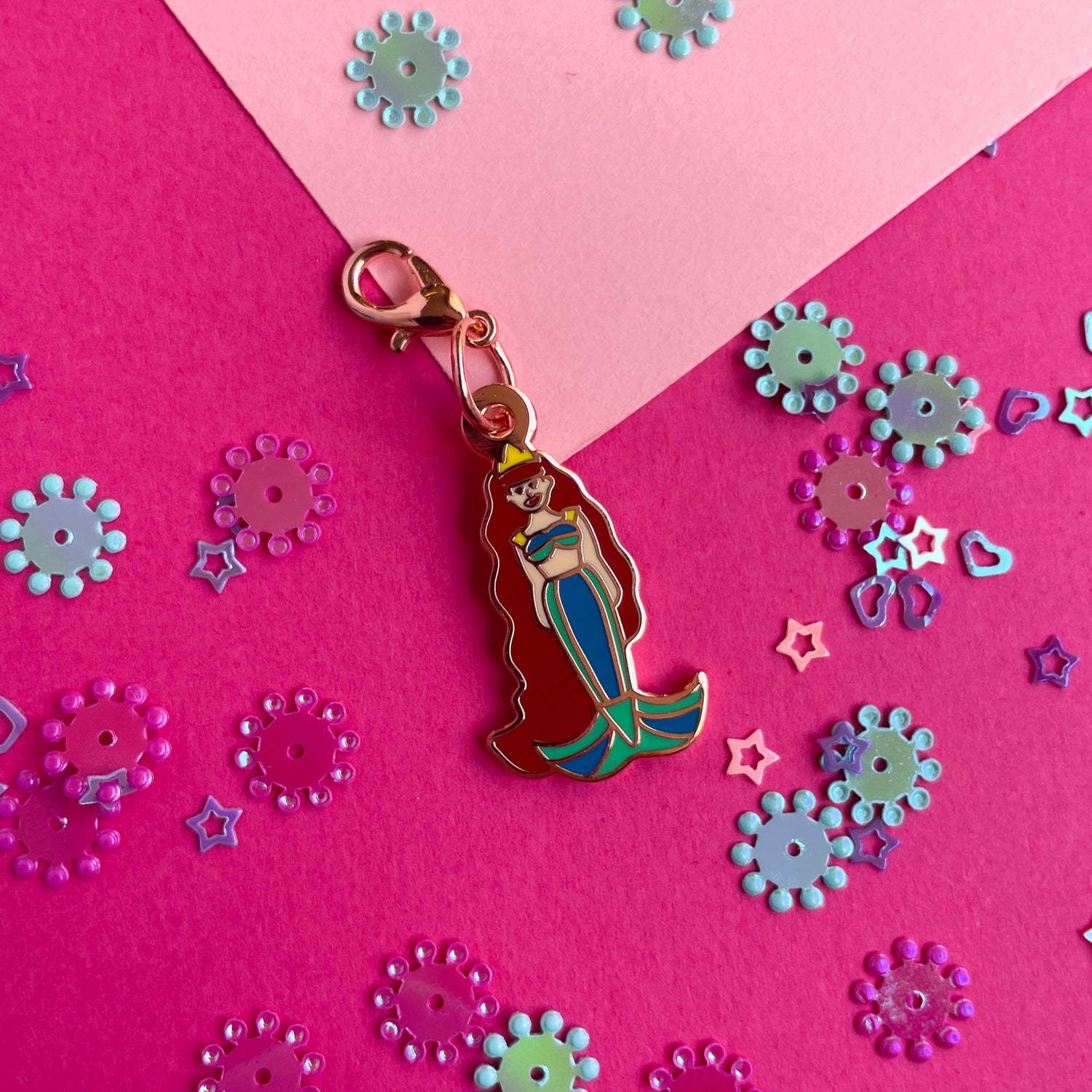 A lobster claw clasp charm shaped like a mermaid with long red hair and a green tail on a pink paper background.