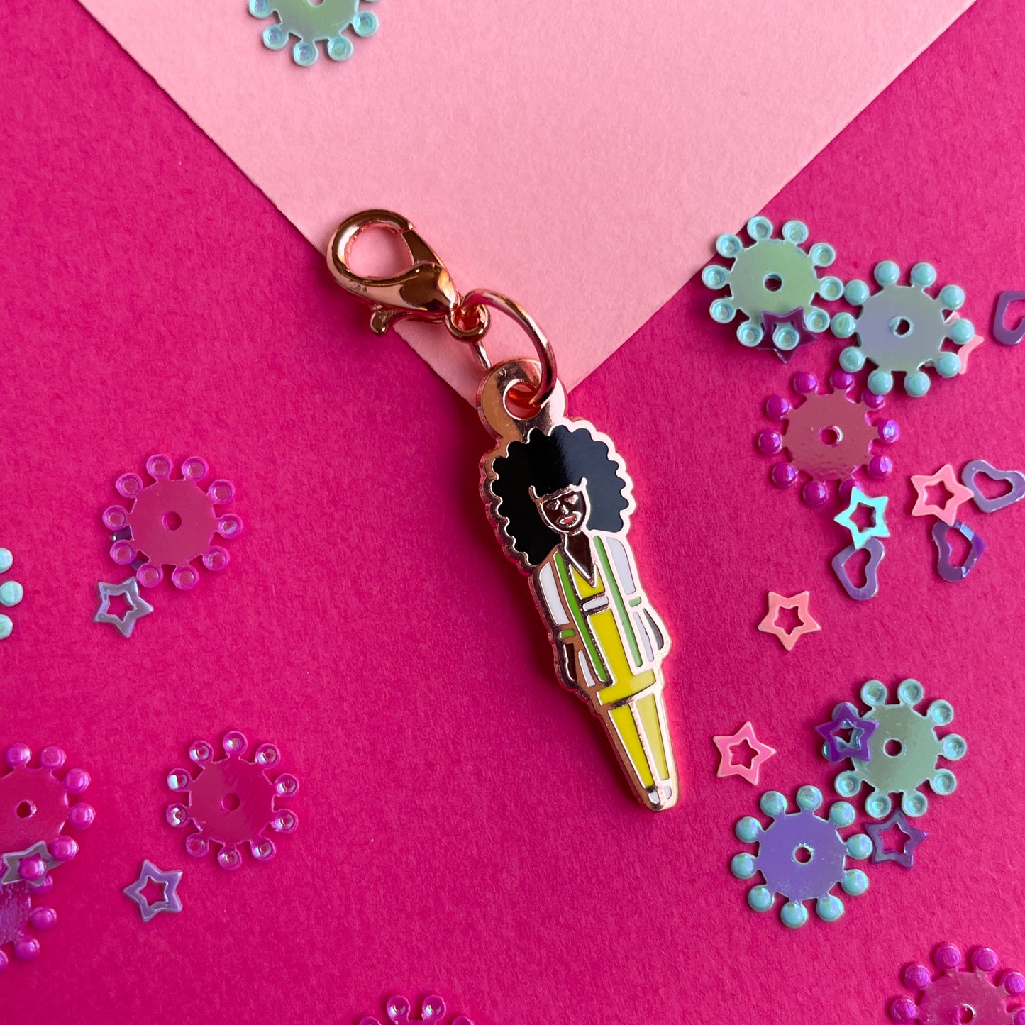 An enamel charm shaped like a barbie doll with dark brown skin and an afro in a yellow suit on a pink paper background.
