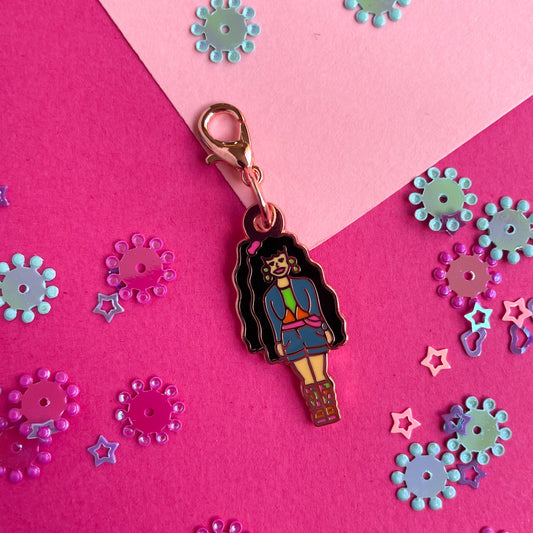 An enamel charm shaped like a barbie with long dark black hair wearing 90s clothes on a pink paper background.