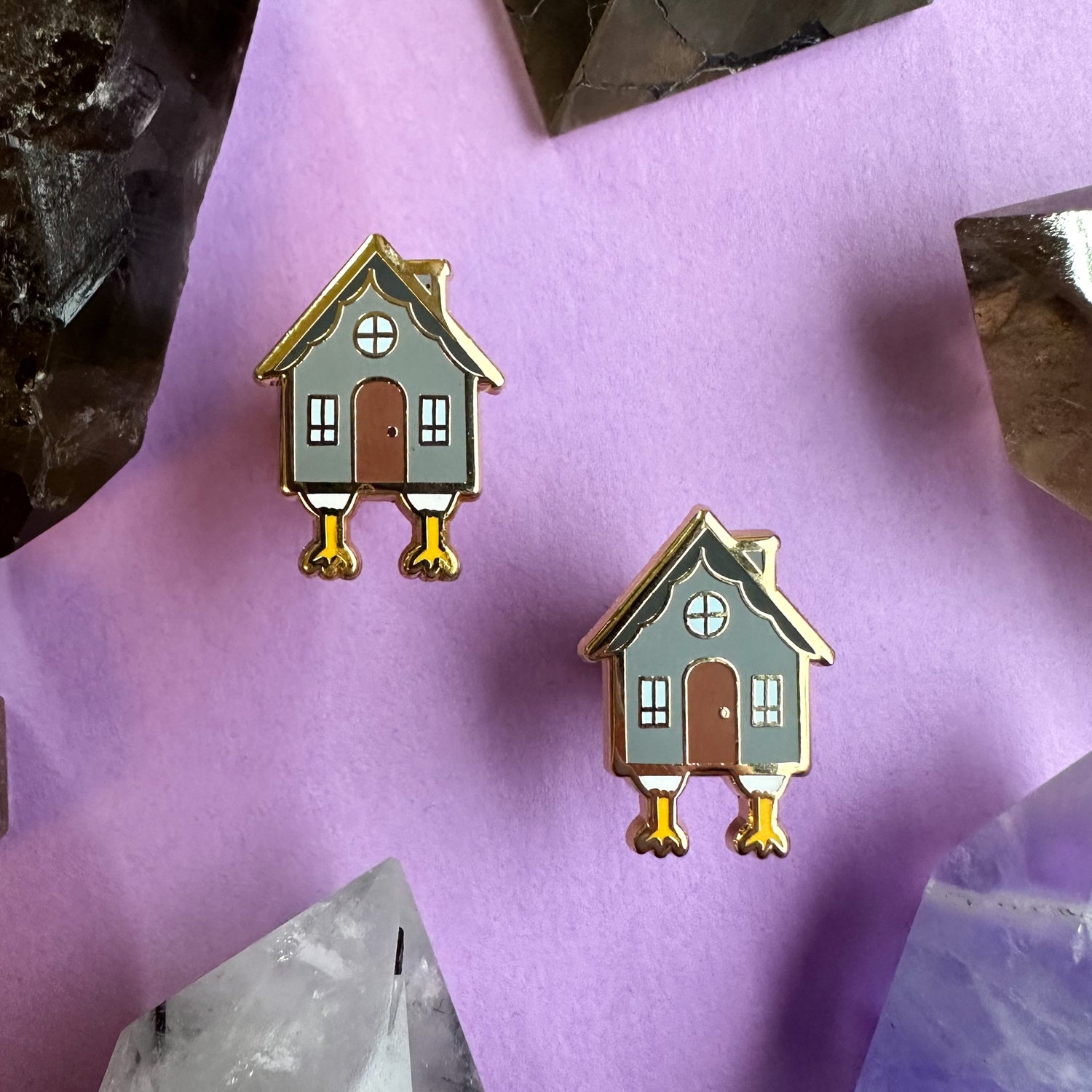 Earrings shaped like Baba Yaga's house on a purple paper background surrounded by crystals 