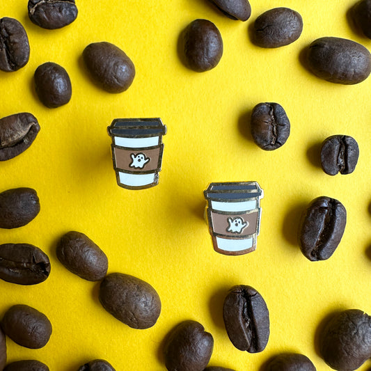 Coffee cup earrings with a ghost on them on a yellow paper background covered in coffee beans