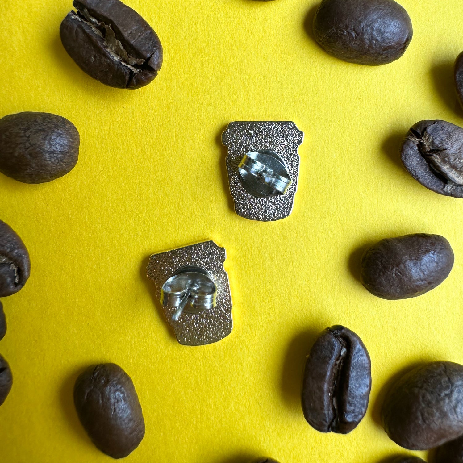The back of the coffee cup earrings with metal butterfly backs on a yellow paper background covered in coffee beans