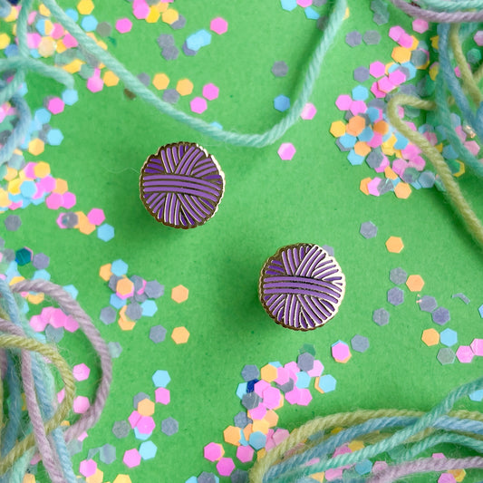 Purple yarn ball earrings on a green paper background covered in yarn and confetti