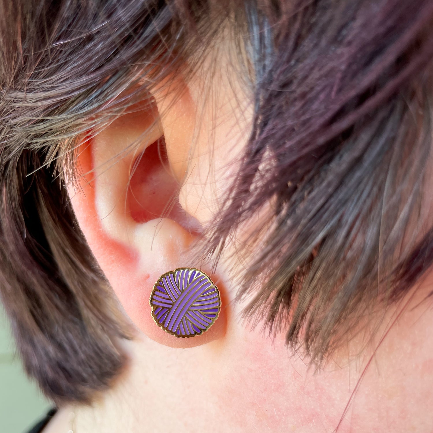 A close up of an ear with short choppy purple hair around it, there is a purple yarn ball earring on the earlobe. 