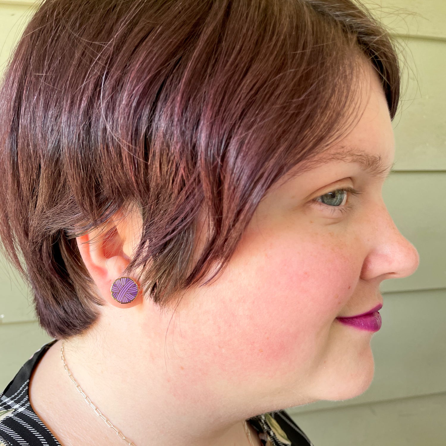 A close up of a woman with a short pixie cut wearing a purple yarn ball shaped post earring. 