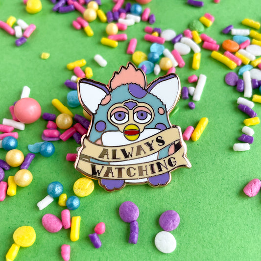 An enamel pin in the shape of a Furby with a tattoo style banner around its body that says "Always Watching". The Furby is light blue with peach and lavender spots. The pin is on a green paper background that is covered with pastel sprinkles