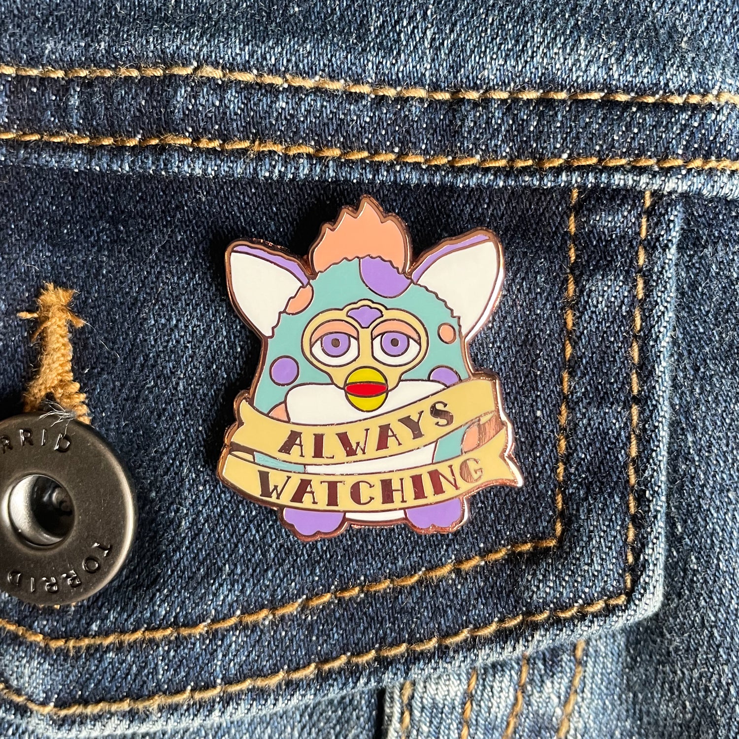 An enamel pin in the shape of a Furby with a tattoo style banner around its body that says "Always Watching". The Furby is light blue with peach and lavender spots. The pin is on a denim pocket.