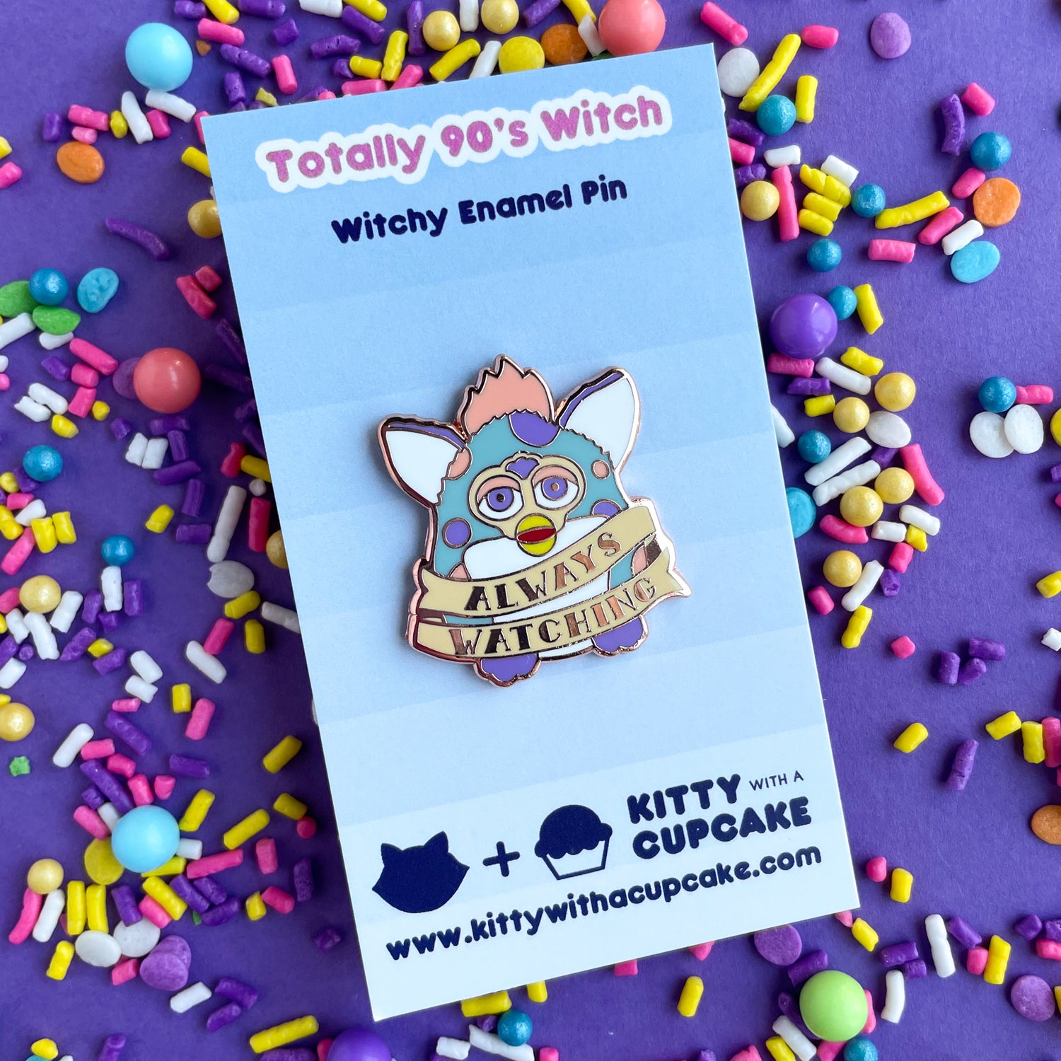 An enamel pin in the shape of a Furby with a tattoo style banner around its body that says "Always Watching". The Furby is light blue with peach and lavender spots. The pin is on a backing card that reads "Totally 90's Witch" on a purple paper background that is covered in sprinkles.