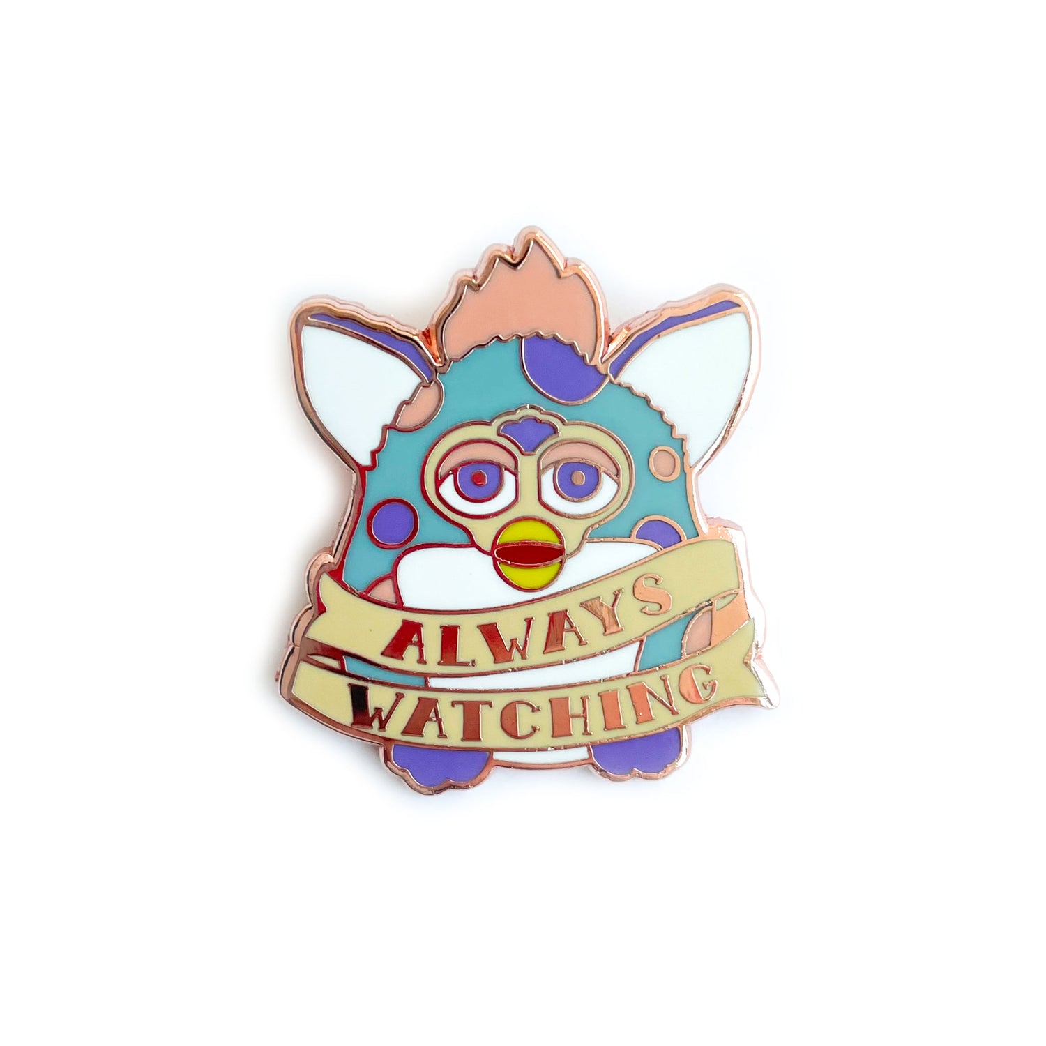 An enamel pin in the shape of a Furby with a tattoo style banner around its body that says "Always Watching". The Furby is light blue with peach and lavender spots. 