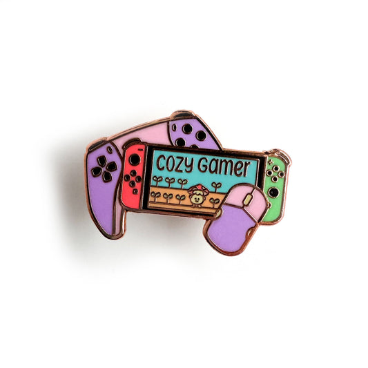 An enamel pin shaped like a Nintendo switch with a mouse and a game controller that has the words "Cozy Gamer" on the screen with a cute mushroom.