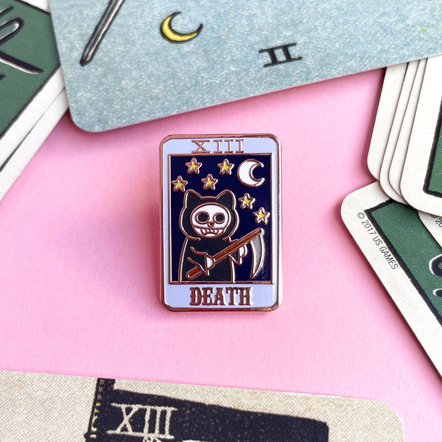 An enamel pin shaped like a tarot card with a grim reaper cat with a night sky on it and the word "Death" surrounded by other tarot cards.