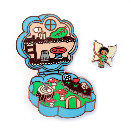 Two enamel pins, one of a small fairy doll in a green dress, the other of a flower compact set up as a fairy dollhouse with mushrooms and flowers.