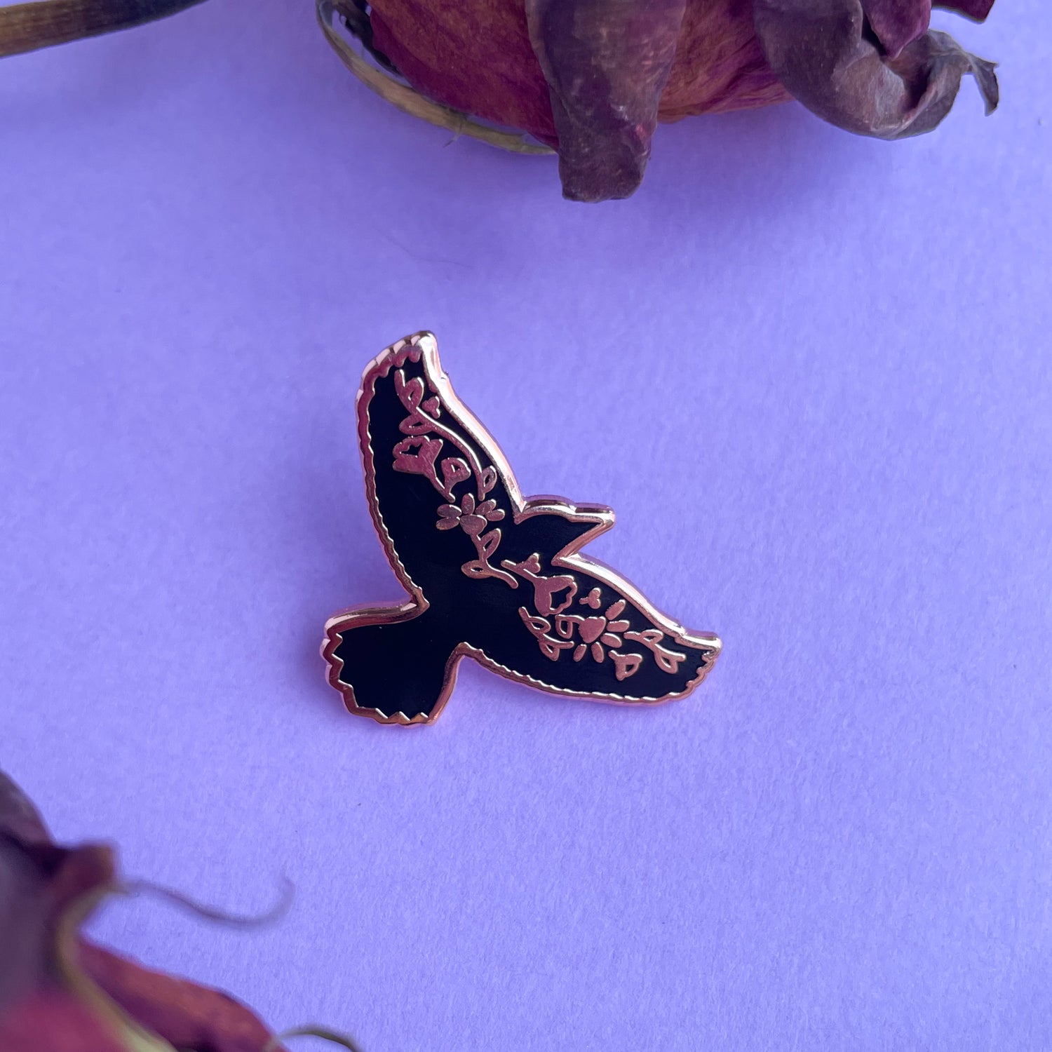 An enamel pin shaped like the silhouette of a raven with a floral pattern along its wings. 
