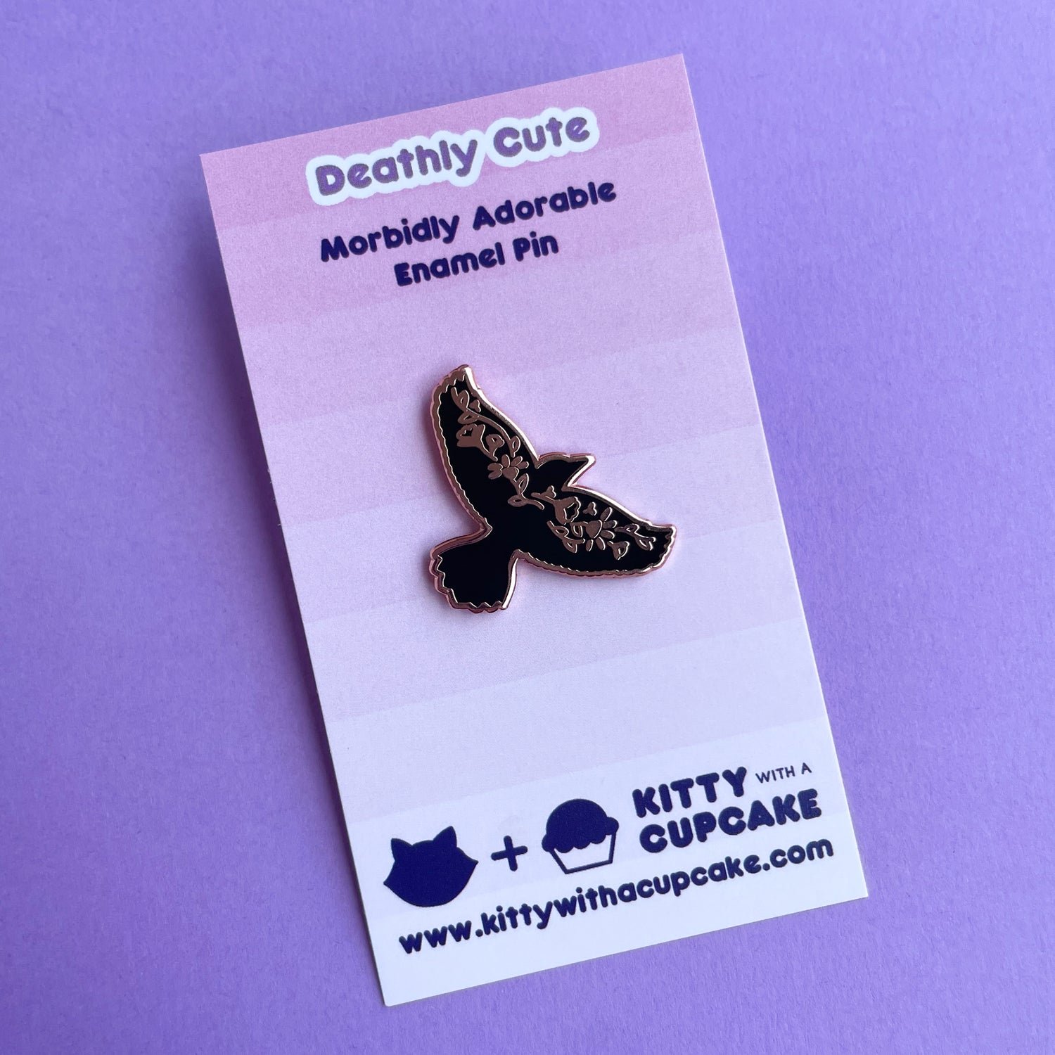 An enamel pin shaped like the silhouette of a raven with its wingspread on a pink card.