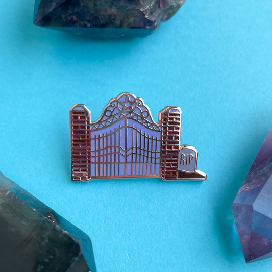 An enamel pin shaped like a cemetery gate on a blue background surrounded by crystals.