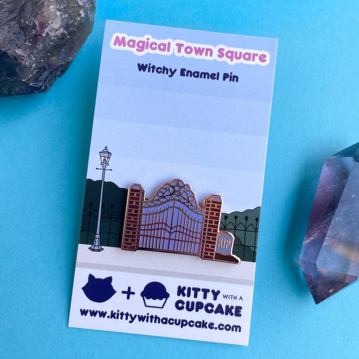 An enamel pin of a cemetery gate on a card with the scene of a town. 