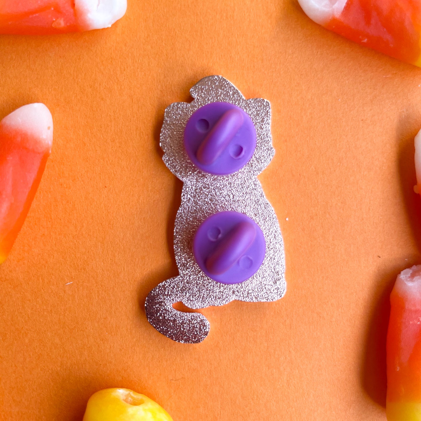 The back of an enamel pin with purple rubber pin backs on an orange background with candy corn.