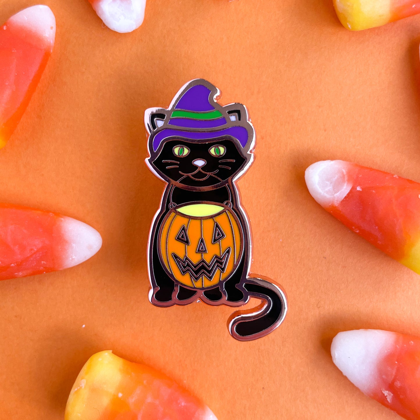 An enamel pin shaped like a black cat wearing purple witch hat holding an orange jack o lantern bucked in its mouth on an orange background with candy corn. 