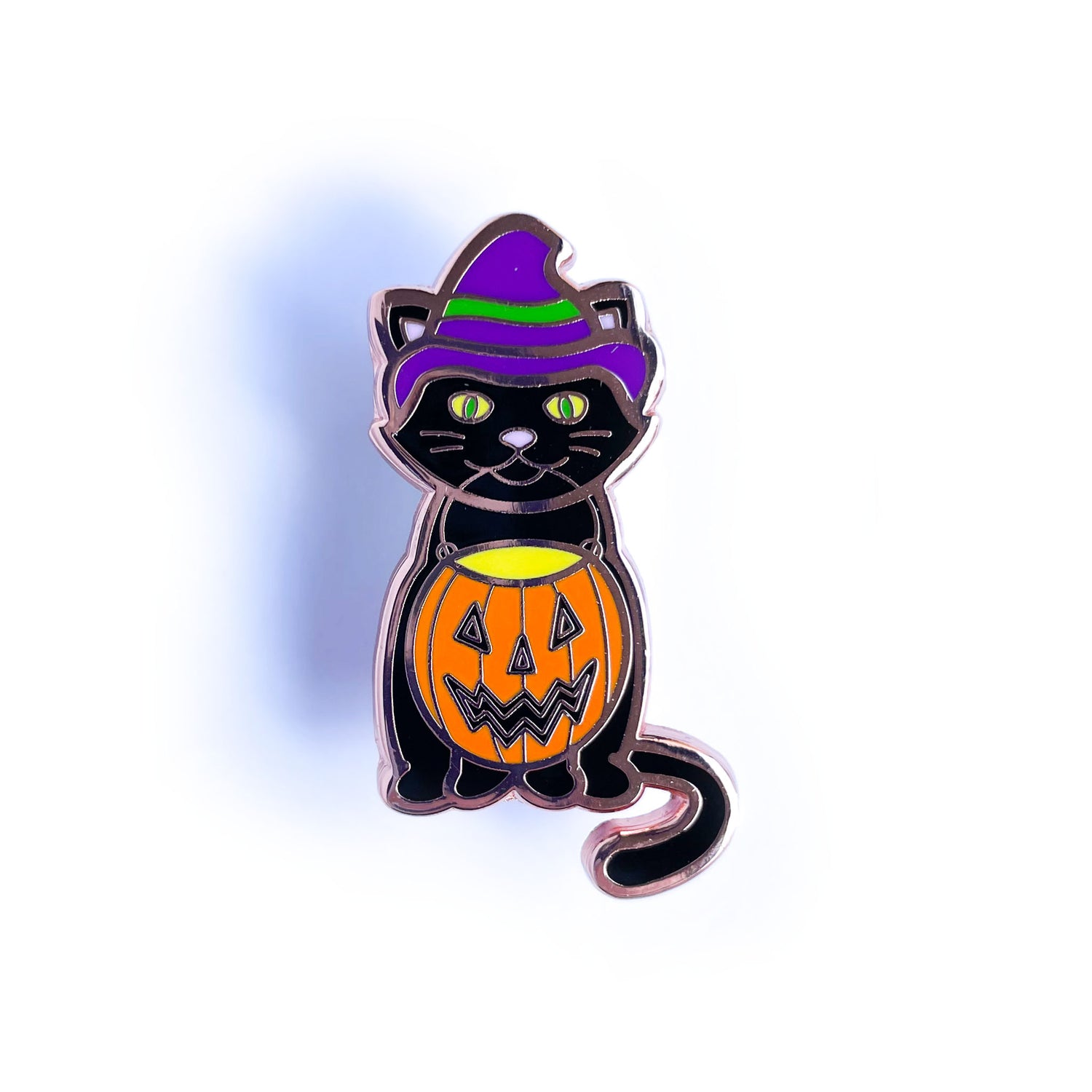 An enamel pin shaped like a black cat wearing purple witch hat holding an orange jack o lantern bucked in its mouth.