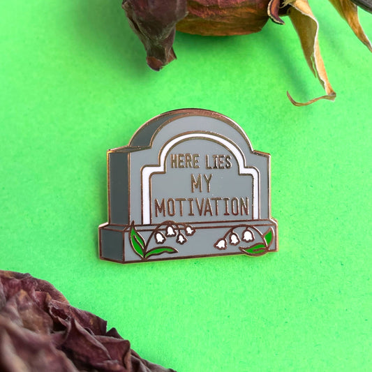 A gravestone shaped pin that reads "Here Lies My Motivation" with lily of the valley flowers in front of it on a green background