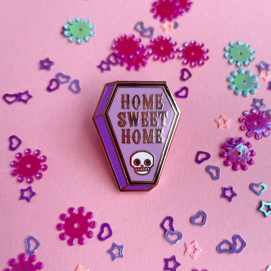 A coffin shaped enamel pin with the words "Home Sweet Home" on it and a skull on a pink background with confetti around it. 